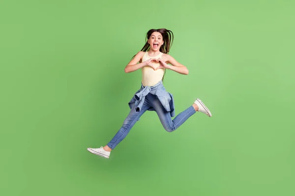 Фотографія повного розміру щасливої веселої збудженої божевільної дівчинки, що стрибає, показуючи знак серця ізольовано на фоні зеленого кольору — стокове фото