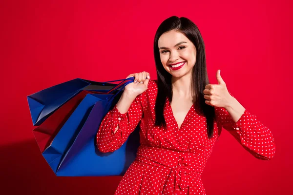 Фотографія радісної брюнетки леді з сумками показують великий палець в сукні червоного кольору ізольовані на яскравому кольоровому фоні — стокове фото