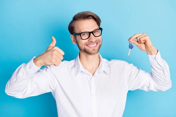 Фото веселого молодого привабливого брюнетки чоловік тримає ключі показують посмішку великого пальця ізольовано на синьому кольорі — стокове фото