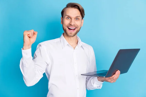 Фото здивованого щасливого переможця молодого чоловіка, який піднімає кулак, тримає ноутбук переможця ізольовано на синьому кольоровому фоні — стокове фото