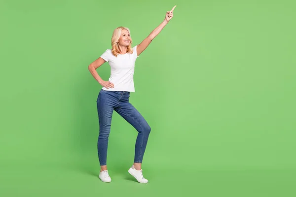 Full size foto van coole blonde dame punt lege ruimte dragen witte t-shirt jeans geïsoleerd op groene kleur achtergrond — Stockfoto