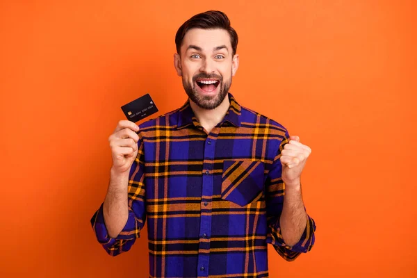 Foto van verrukt knappe jongen vuist omhoog open mond houden debetkaart vieren geïsoleerd op oranje kleur achtergrond — Stockfoto