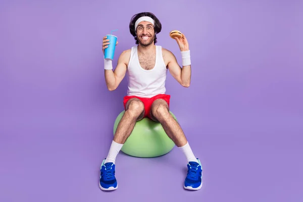 Full size of young happy cheerful positive funky man in glasses sit ball eat hamburger drink cola isolated on purple color background — Stock Photo, Image