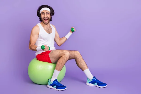 Full size photo of funky happy young man hold dumbbells sit green fit ball smile isolated on purple color background — Stock Photo, Image