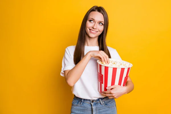 Photo portrait of curious girl with brown hair eating pop corn looking at blank space smiling isolated vibrant yellow color background — Stock Photo, Image