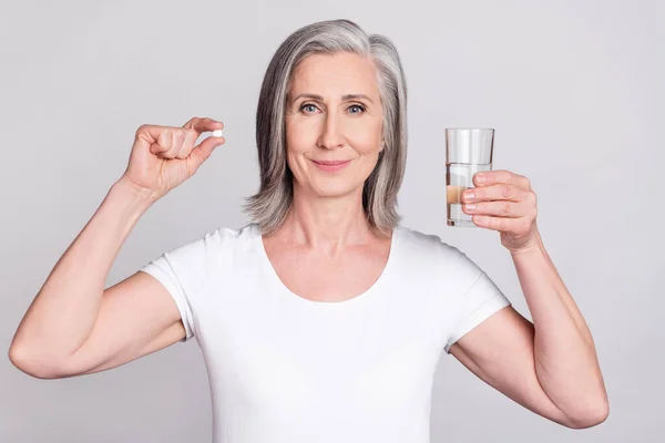 Foto da mulher idosa feliz sorriso positivo segurar drogas de água de vidro analgésico isolado sobre fundo de cor cinza — Fotografia de Stock