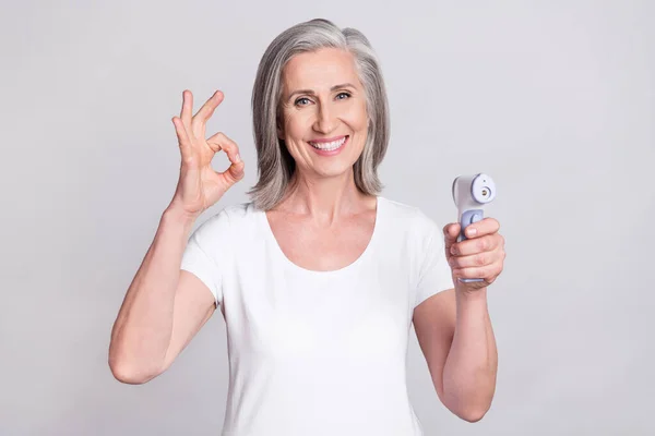 Foto da mulher idosa feliz sorriso positivo mostrar tudo bem sinal digitalizar temperatura termômetro isolado sobre fundo de cor cinza — Fotografia de Stock