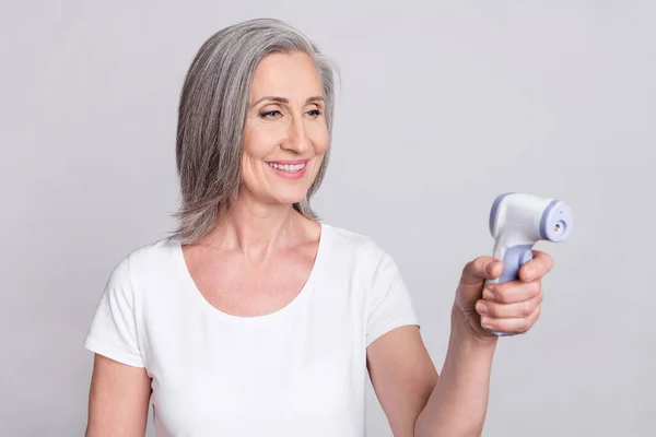 Foto de senhora idosa positiva mantenha o termômetro sem contato use camiseta branca isolada no fundo de cor cinza — Fotografia de Stock