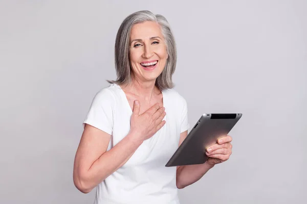 Foto de linda hora divertida señora madura celebrar tableta risa usar camiseta blanca aislada sobre fondo de color gris —  Fotos de Stock