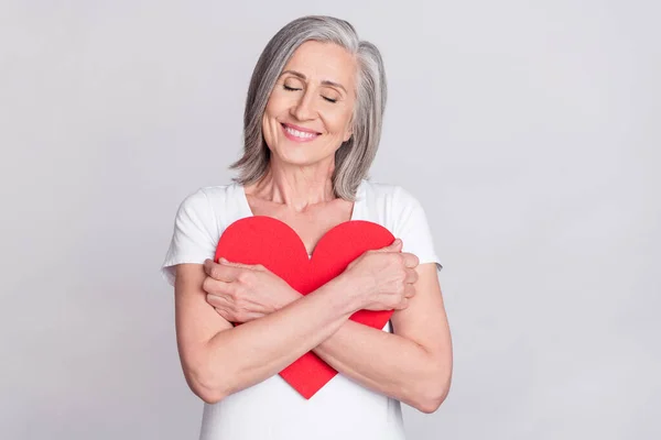 Foto de mulher adorável idade sonhadora vestida roupa branca sorrindo abraçando grande coração vermelho isolado fundo de cor cinza — Fotografia de Stock