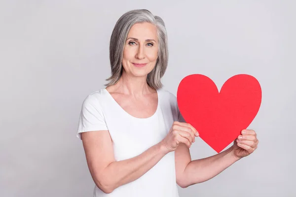 Fénykép az idős nő boldog pozitív mosoly hold piros papír szív kártya szerelem romantikus elszigetelt felett szürke színű háttér — Stock Fotó