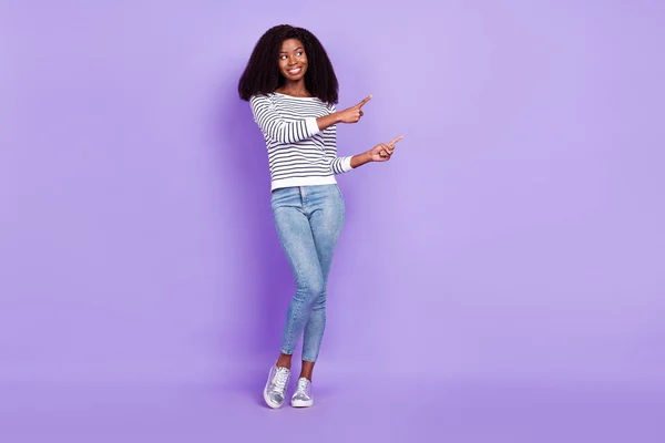 Full body foto van optimistische jonge dame punt lege ruimte slijtage gestreepte pullover jeans geïsoleerd op violette kleur achtergrond — Stockfoto