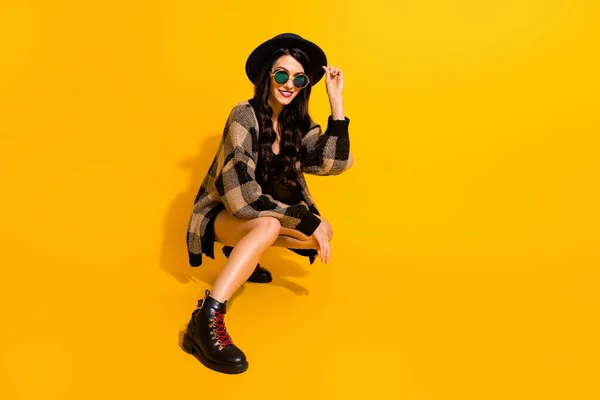 Foto do hipster jovem senhora sentar olhar câmera desgaste óculos retro chapéu xadrez casaco isolado cor amarela fundo — Fotografia de Stock