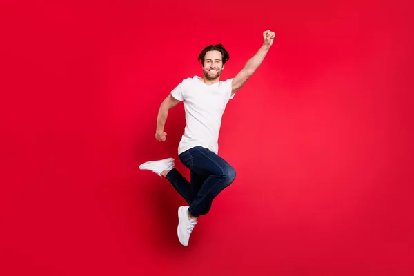 Ganzkörper-Foto von jungen braunen Frisur Mann springen tragen weißes T-Shirt Jeans isoliert auf rotem Hintergrund — Stockfoto