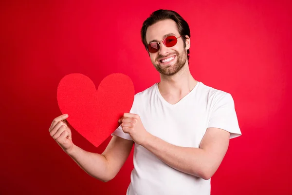 Foto de belo jovem homem penteado marrom segurar papel coração desgaste óculos branco t-shirt isolado na cor vermelha fundo — Fotografia de Stock
