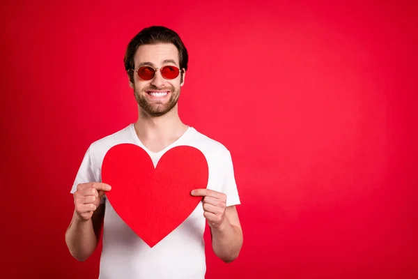 Fotografie sladké mladé hnědé vlasy muž držet papír srdce vypadat prázdný prostor nosit brýle bílá tričko izolované na červeném pozadí — Stock fotografie