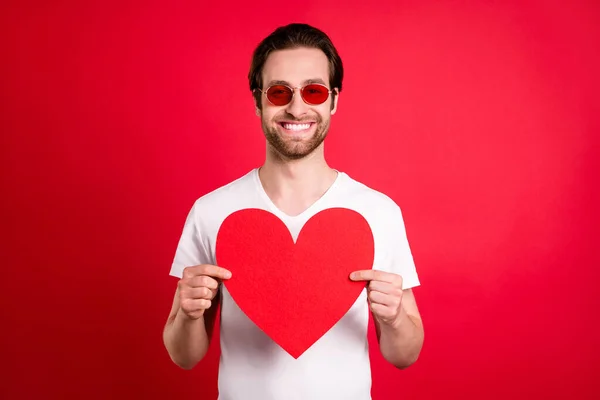 Foto de paquera jovem marrom penteado homem segurar papel coração desgaste óculos branco t-shirt isolado na cor vermelha fundo — Fotografia de Stock