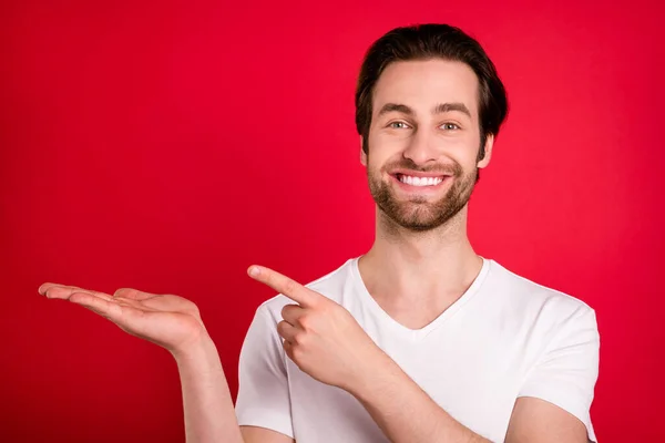 Foto von optimistischen Millennial braune Frisur Mann halten Punkt leeren Raum tragen weißes T-Shirt isoliert auf rotem Hintergrund — Stockfoto