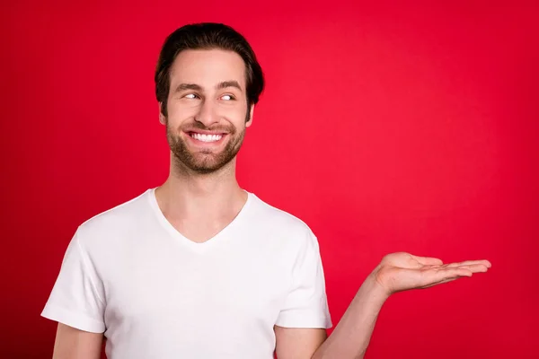 Foto av roliga millennial brun frisyr man hålla handen ser tomt utrymme slitage vit t-shirt isolerad på levande röd färg bakgrund — Stockfoto