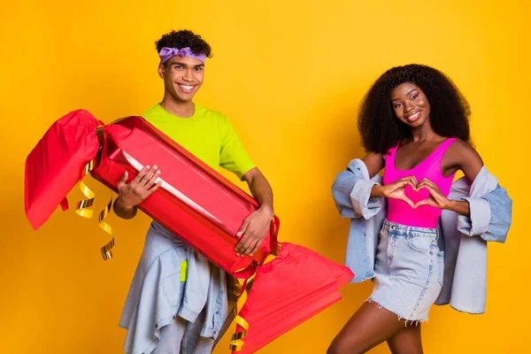 Foto de pareja joven coqueta brillante vestido traje de mezclilla que sostiene enormes dulces que muestran los dedos corazón aislado color amarillo fondo —  Fotos de Stock