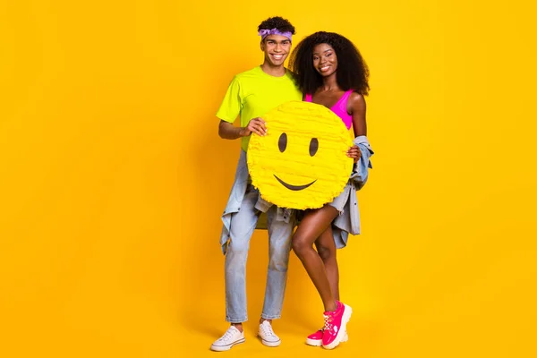 Foto av söta glänsande unga par klädd denim outfit hålla stora smiley tecken kram omfamna isolerad gul färg bakgrund — Stockfoto