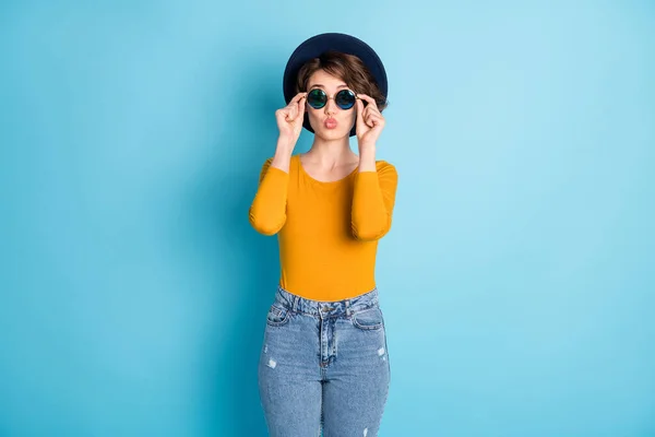 Фото оптимистичной брюнетки леди сенсорные очки удар поцелуй ношение шапки желтая рубашка изолированы в ярко-голубой цвет фона — стоковое фото