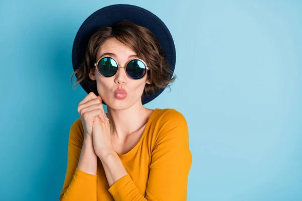 Foto di ottimista bruna signora tenere per mano colpo bacio indossare cappello occhiali camicia gialla isolato in vivido sfondo di colore blu — Foto Stock