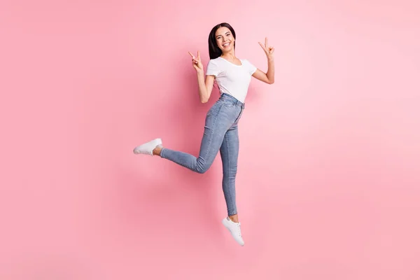 Full storlek profil foto av optimistiska brunett dam hoppa visar v-tecken slitage t-shirt jeans sneakers isolerade på pastell rosa färg bakgrund — Stockfoto