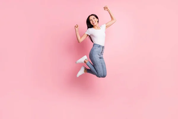 Foto de perfil de tamaño completo de la mujer morena optimista salto manos puños usan zapatillas vaqueros camiseta aislada sobre fondo de color rosa pastel —  Fotos de Stock