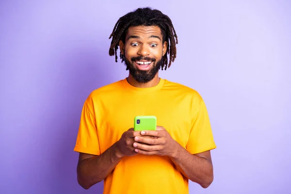 Retrato de bonito peinado morena impresionado chico mira teléfono desgaste naranja camiseta aislada sobre fondo de color lila — Foto de Stock