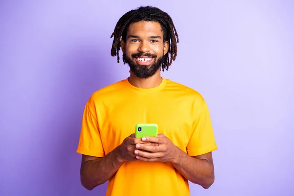 Güzel, iyimser esmer adamın portresi telefon tut turuncu t-shirt giy parlak leylak rengi arka planda izole edilmiş — Stok fotoğraf