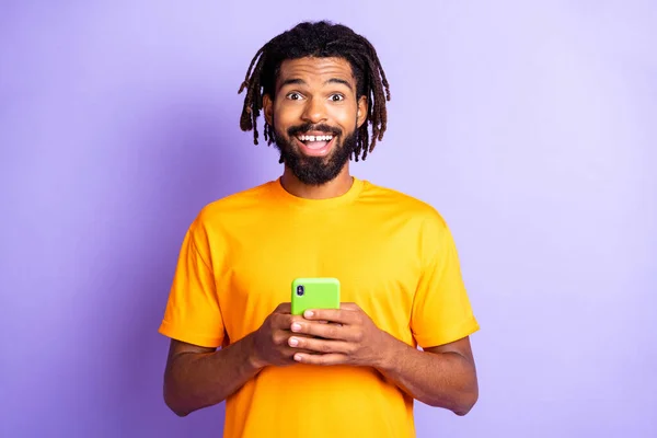 Portret van mooie optimistische brunet guy type telefoon dragen oranje t-shirt geïsoleerd op levendige lila kleur achtergrond — Stockfoto