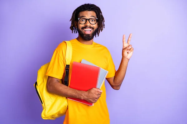 Портрет симпатичного впечатлённого брюнет парень держит книги показывают V-знак носить очки мешок оранжевая футболка изолированы на ярком сиреневом фоне — стоковое фото