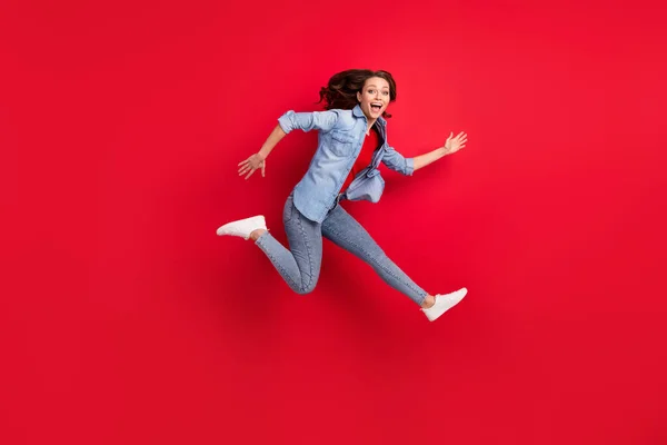 Foto de adorável animado jovem mulher usar jeans camisa sorrindo saltando alto apressando isolado fundo cor vermelha — Fotografia de Stock