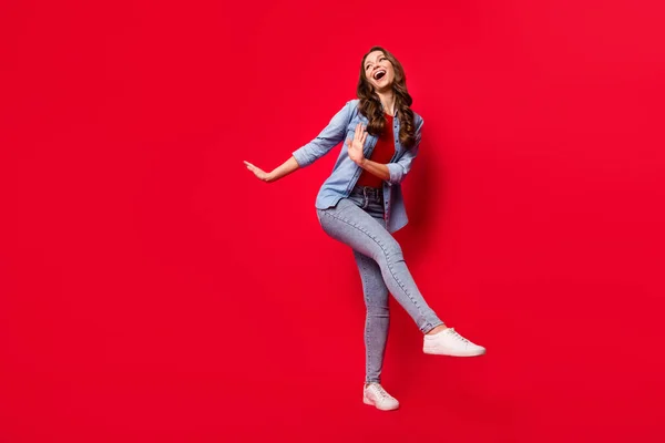 Foto de excitado divertido joven vestido traje de mezclilla caminando bailando sonriendo buscando espacio vacío aislado color rojo fondo —  Fotos de Stock