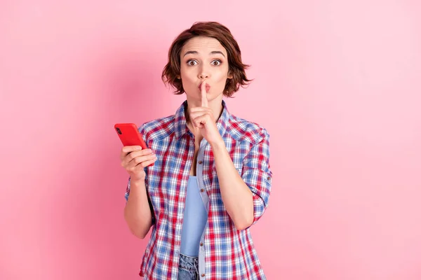 Foto de adorable joven mujer usar camisa a cuadros que sostiene gadget moderno pidiendo no contar secretos aislado color rosa fondo — Foto de Stock