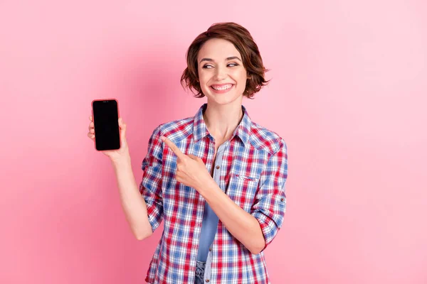 Foto de bonito brilhante jovem senhora vestida camisa xadrez apontando segurando dispositivo moderno olhando espaço vazio isolado cor de fundo rosa — Fotografia de Stock