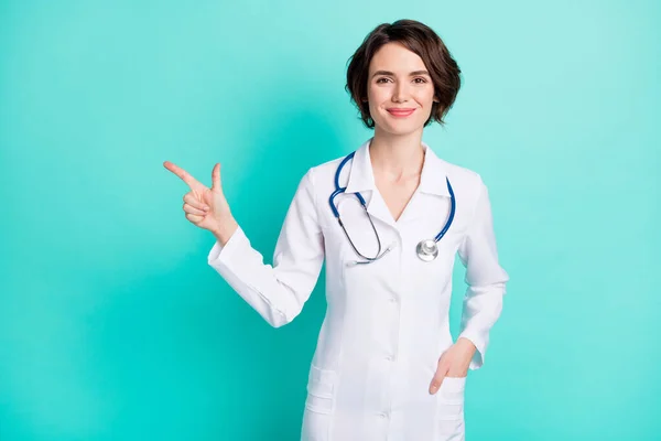 Fénykép a fiatal nő orvos boldog mosoly jelzi ujj üres tér hirdetés ajánlom válassza elszigetelt felett teal színes háttér — Stock Fotó