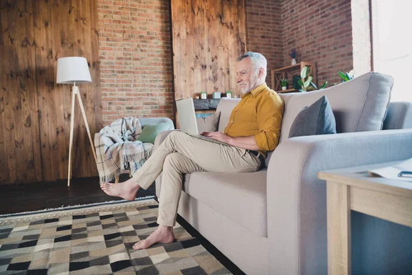 Πλήρες προφίλ του σώματος πορτρέτο του χαρούμενου ηλικιωμένου άνδρα κάθονται στον καναπέ ματιά φορητό υπολογιστή toothy χαμόγελο δακτυλογράφηση μασάζ στο σπίτι σε εσωτερικούς χώρους — Φωτογραφία Αρχείου