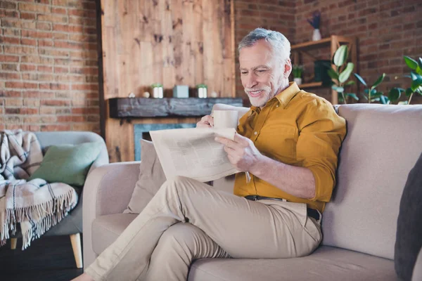 Photo of positive aged man sit on couch holding coffee reading interested newspaper enjoy home indoors — Stock Photo, Image