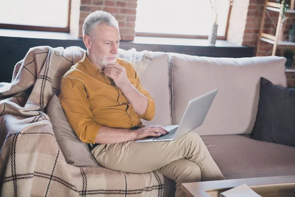 Photo of minded poważne człowiek siedzieć na kanapie ramię w brodzie wygląd zainteresowany koncentruje laptop myśląc w domu w pomieszczeniach — Zdjęcie stockowe