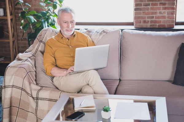 Portret pozytywnego starca siedzieć na kanapie trzymać wygląd używać laptopa oglądać filmy mają dobry nastrój pracy z domu w pomieszczeniach — Zdjęcie stockowe