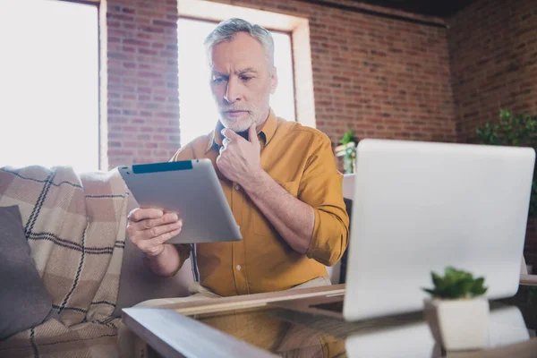 Photo of serious minded aged person sit on couch hand on chin look interested tablet thought working from home indoors — Stock Photo, Image