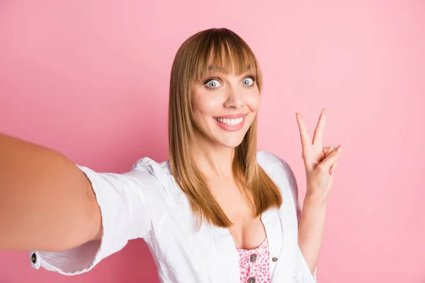 Foto ritratto blogger femminile scattare selfie dimostrando gesto v-segno sorridente in camicia bianca isolato su sfondo rosa pastello — Foto Stock