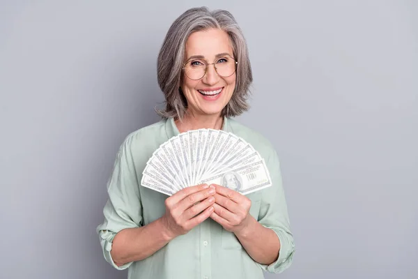Foto di felice eccitata allegra nonna positiva in occhiali tenere soldi dollari isolati su sfondo di colore grigio — Foto Stock