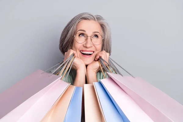 Fénykép érett boldog álmodozó nő szemüvegben néz copyspace hold bevásárló táskák elszigetelt szürke színű háttér — Stock Fotó