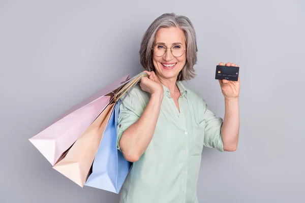 Foto de feliz sorrindo alegre vovó mostrar cartão de crédito segurar sacos de compras shopaholic isolado no fundo de cor cinza — Fotografia de Stock