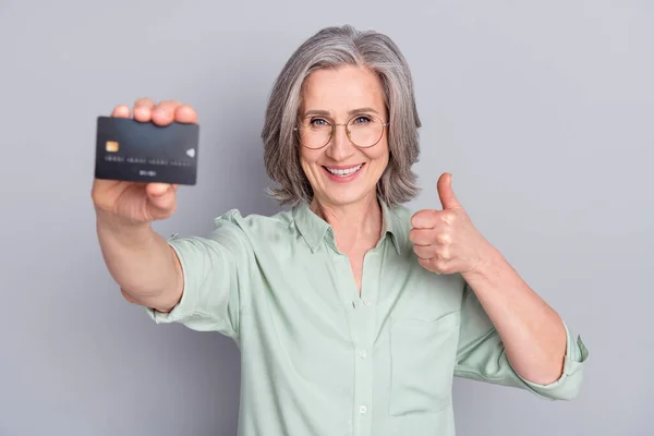 Retrato de idoso alegre atraente aposentado aposentado segurando cartão bancário mostrando polegar isolado sobre fundo cor pastel cinza — Fotografia de Stock