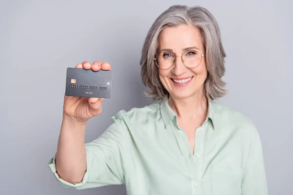 Foto de feliz sorrindo positivo mulher muito madura em óculos mostrar-lhe cartão de crédito isolado em fundo de cor cinza — Fotografia de Stock