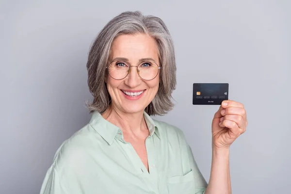 Foto de feliz sorrindo bela mulher madura em óculos mostrando cartão de crédito isolado em fundo de cor cinza — Fotografia de Stock